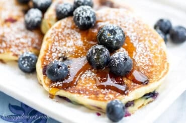A closeup of a platter of pancakes with blueberries and syrup on top.