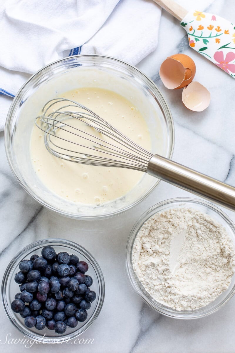 A bowl of pancake batter with a whisk and a bowl of blueberries.