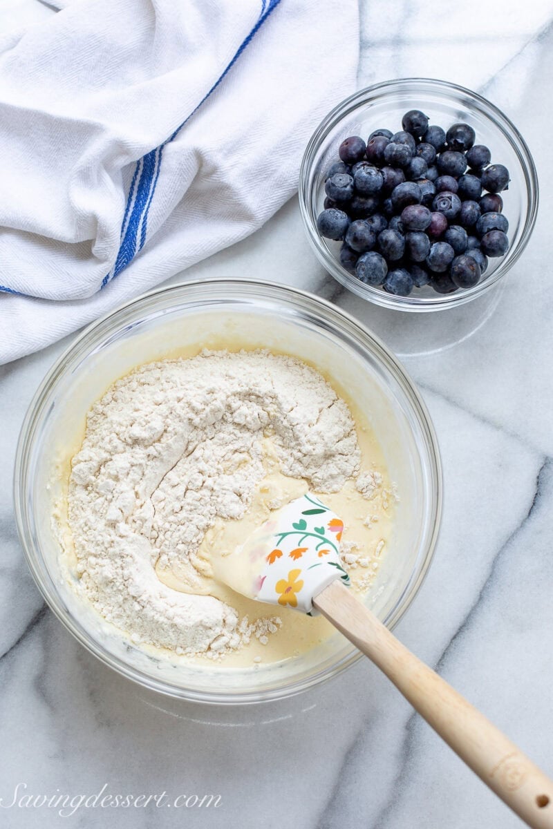 A bowl of pancake batter with flour being folded in with blueberries on the side.