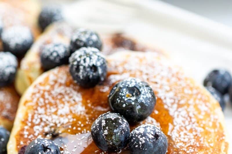 A platter of pancakes topped with fresh blueberries, powdered sugar and a drizzle of syrup.
