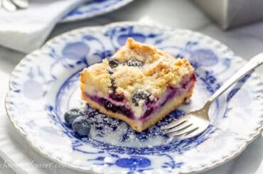 A closeup photo of a blueberry cheesecake bar on a white and blue plate with a small fork on the side.