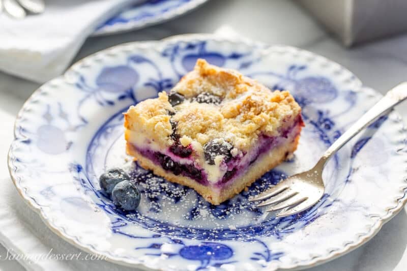 A closeup photo of a blueberry cheesecake bar on a white and blue plate with a small fork on the side.