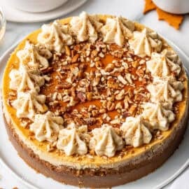 A whole maple cheesecake on a platter with maple buttercream swirls on top with maple syrup and pecans.