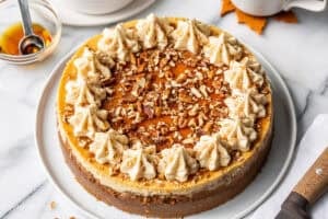 A whole maple cheesecake on a platter with maple buttercream swirls on top with maple syrup and pecans.