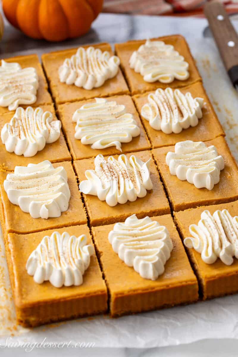 A side view of a pan of pumpkin cheesecake bars cut into squares each topped with swirls of whipped cream.