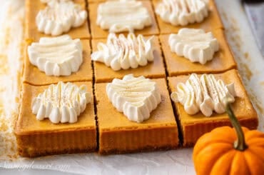 A pan of pumpkin cheesecake bars cut into squares with a mini pumpkin close by.