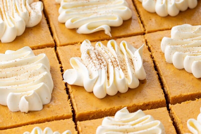 Pumpkin cheesecake bars cut into squares topped with swirls of whipped cream.