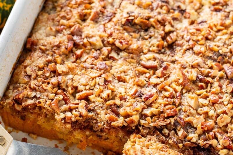 An overhead view of a baking pan with pumpkin dump cake topped with pecans and a spatula scooping out a piece.