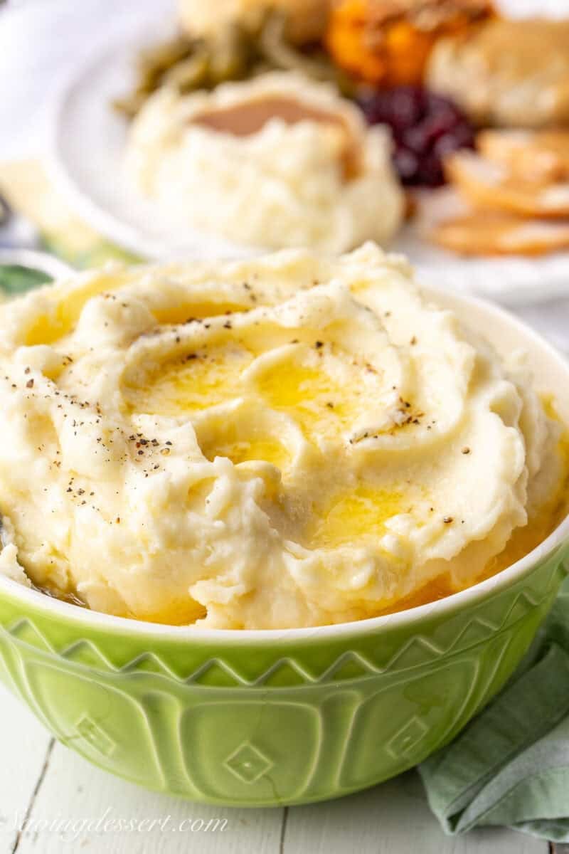 A side view of a bowl of creamy mashed potatoes with melted butter on top and a plate of Thanksgiving favorites in the background.