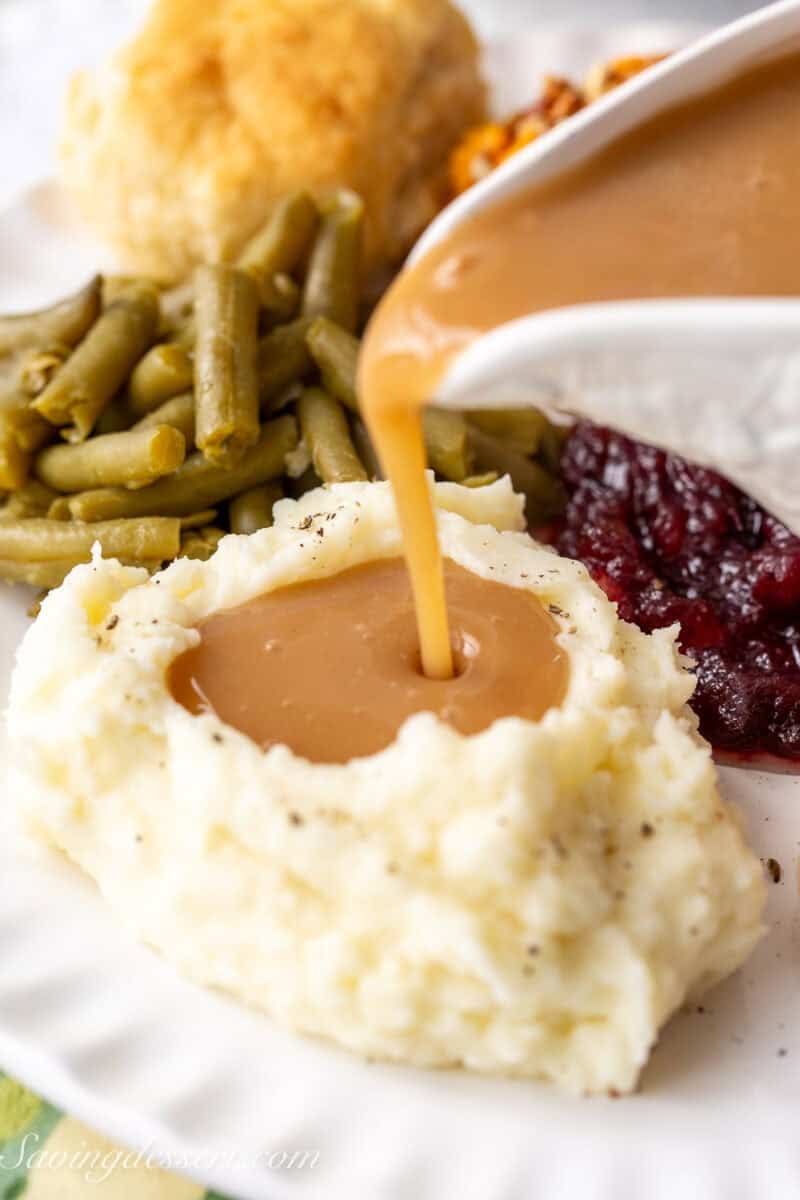 A mound of mashed potatoes on a plate with gravy being poured onto the potatoes.