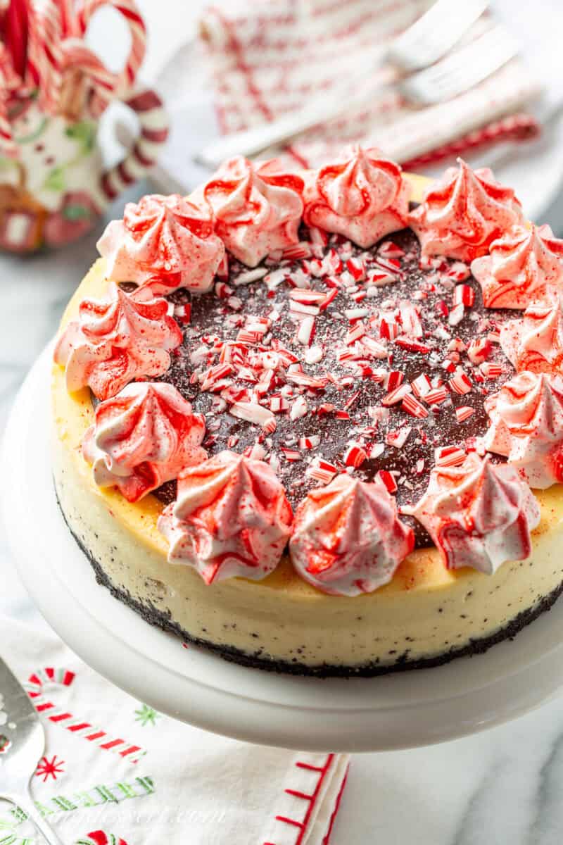 Peppermint cheesecake on a cake stand decorated with whipped cream swirls, chocolate ganache and crushed peppermints.
