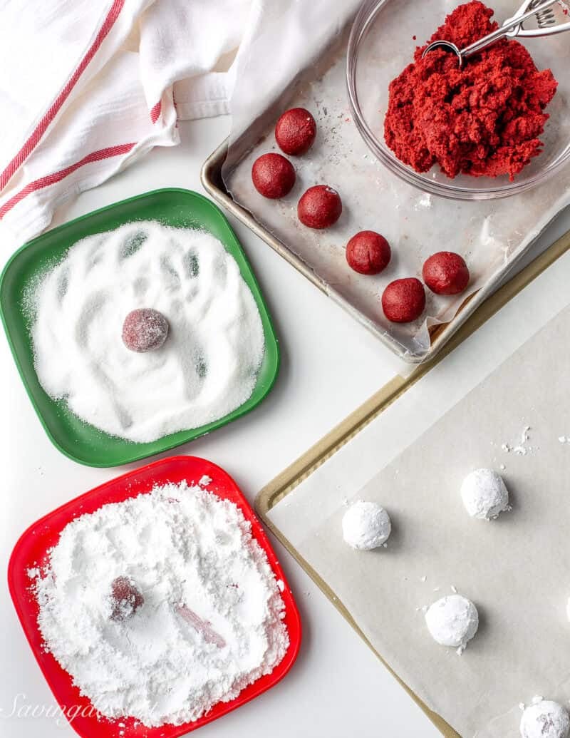 Red velvet cookie dough balls being rolling in granulated and powdered sugar before baking.