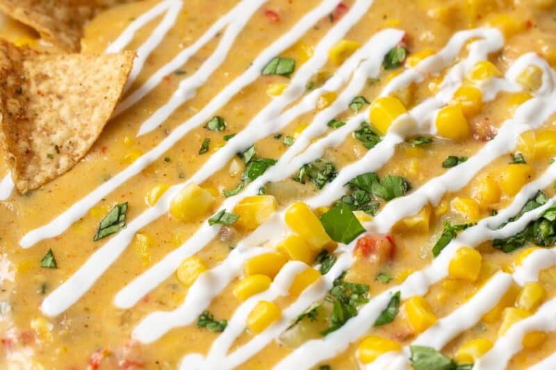 A side view of a bowl of Mexican Street Corn drizzled with Mexican crema, limes and tortilla chips.