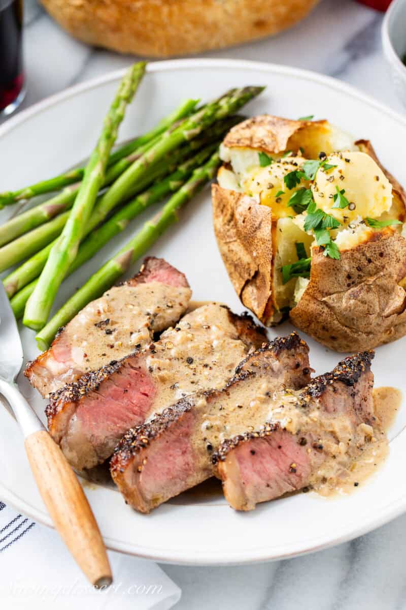 A closeup of a plate with sliced steak topped with a brandy cream sauce and served with a baked potato and asparagus.