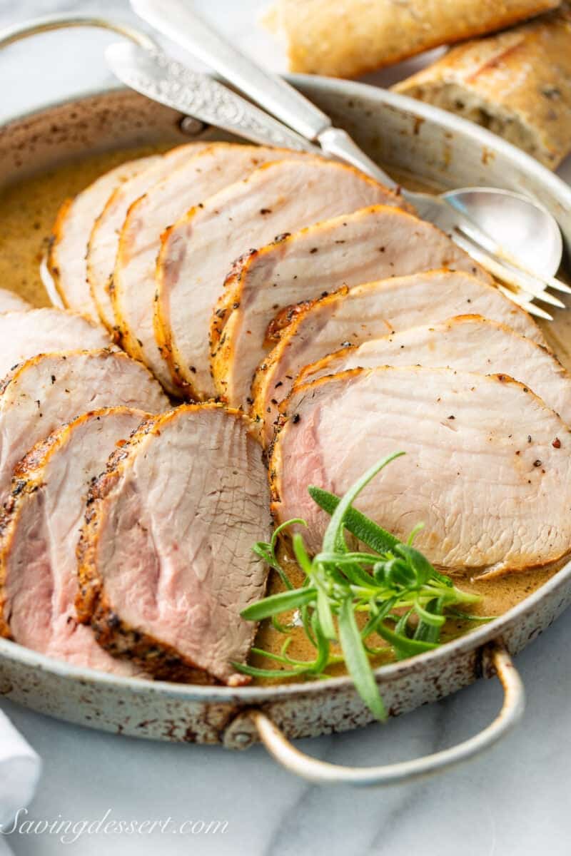 A shallow roasting pan with sliced pork loin in a sauce with fresh rosemary garnish.
