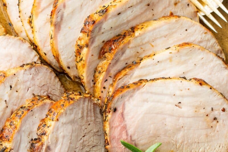 A closeup of a roasting pan filled with sliced pork loin in a sauce with a rosemary sprig as garnish.