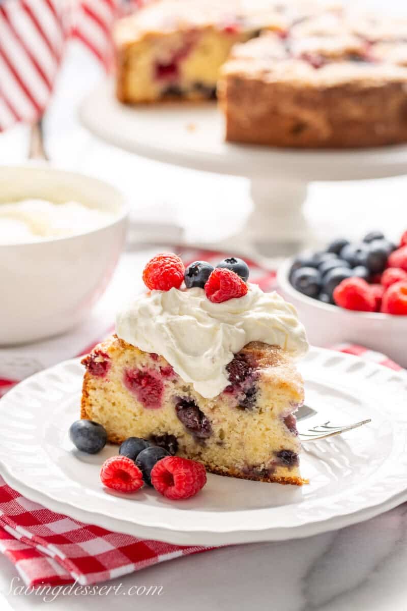 A slice of a berry filled cake with a big dollop of cream on top with raspberries and blueberries as a garnish.