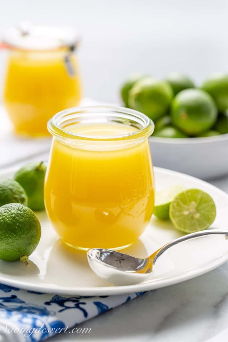 A side view of a clear jar of key lime curd with sliced key limes on a plate.