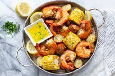 A baking pan loaded with cooked shrimp, potatoes, corn and sausage with melted butter for dipping.